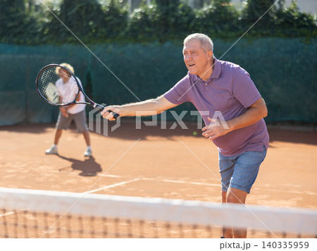 Active aged man hits tennis ball with racket in front of players in court. Two sportive men enjoying doubles match Active aged man hits tennis ball with racket in front of players in court. Two sportive men enjoying doubles match 140335959