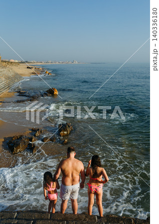 Rear View of Father and Two Daughters Standing by Ocean Waves on Rocky Beach at Sunset 140336180