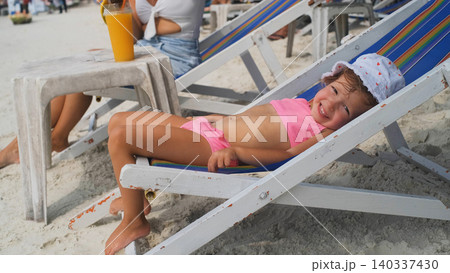 Cute little girl in pink swimsuit lying and relaxing on a beach lounge chair on a sunny tropical beach. Childhood vacation concept Cute little girl in pink swimsuit lying and relaxing on a beach lounge chair on a sunny tropical beach. Childhood vacation concept 140337430