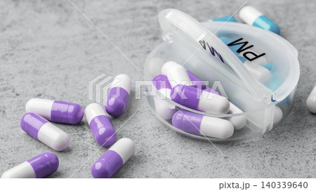 Purple and white capsule pills scattered around with opened daily pill box on a grey concrete surface Purple and white capsule pills scattered around with opened daily pill box on a grey concrete surface 140339640