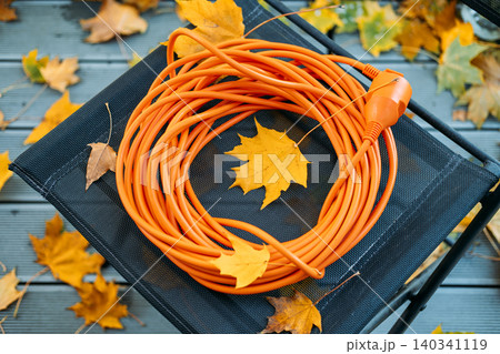 orange extension cord rests on mesh folding chair surrounded by leaves on balcony deck. Blackout preparation solutions, grid resilience autumn, emergency power connection, Backup electricity setup. orange extension cord rests on mesh folding chair surrounded by leaves on balcony deck. Blackout preparation solutions, grid resilience autumn, emergency power connection, Backup electricity setup. 140341119
