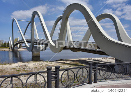 A modern wavy pedestrian bridge spans a calm river at the waterfront. Contemporary architectural arcs create a scenic riverside landmark. A modern wavy pedestrian bridge spans a calm river at the waterfront. Contemporary architectural arcs create a scenic riverside landmark. 140342234