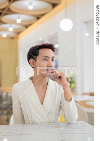 Vertical portrait of thoughtful non-binary gay Thai man looking away at a cafe table 140345396