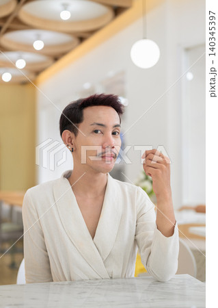 Vertical portrait of non-binary gay Thai man looking at camera sitting at a modern cafe table 140345397