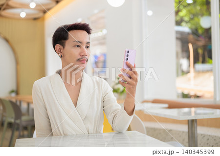 Non-binary gay Thai man looking at smartphone screen while sitting at a modern cafe table Non-binary gay Thai man looking at smartphone screen while sitting at a modern cafe table 140345399