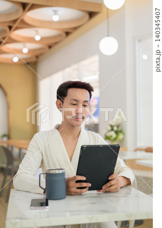 Vertical portrait of non-binary gay Thai man using digital tablet at a modern cafe table Vertical portrait of non-binary gay Thai man using digital tablet at a modern cafe table 140345407