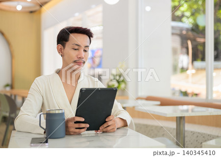 Non-binary gay Thai man reading on digital tablet sitting at modern cafe table Non-binary gay Thai man reading on digital tablet sitting at modern cafe table 140345410