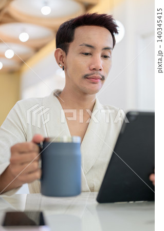 Vertical close up portrait of non-binary gay Thai man holding coffee mug while looking at tablet screen Vertical close up portrait of non-binary gay Thai man holding coffee mug while looking at tablet screen 140345415