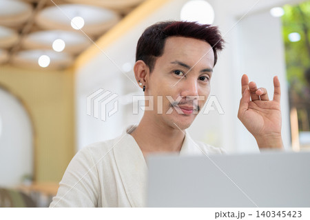 Portrait of smiling non-binary gay Thai man making I love you hand gesture during laptop video call Portrait of smiling non-binary gay Thai man making I love you hand gesture during laptop video call 140345423