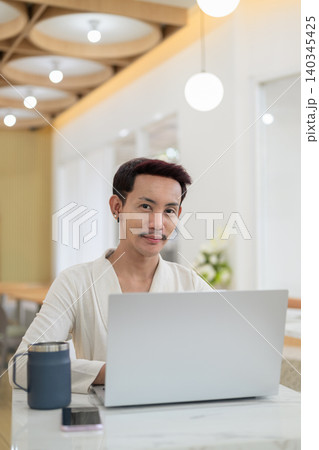 Vertical portrait of non-binary gay Thai man smiling at camera while working on laptop at cafe Vertical portrait of non-binary gay Thai man smiling at camera while working on laptop at cafe 140345425