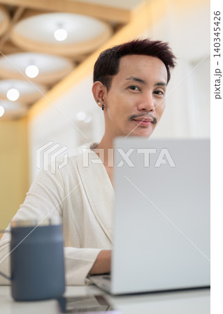 Vertical close up portrait of non-binary gay Thai man smiling over laptop screen at cafe table Vertical close up portrait of non-binary gay Thai man smiling over laptop screen at cafe table 140345426
