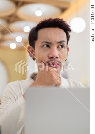 Extreme close up portrait of thoughtful non-binary gay Thai man looking away with hand on chin behind laptop Extreme close up portrait of thoughtful non-binary gay Thai man looking away with hand on chin behind laptop 140345431