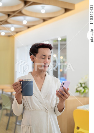 Vertical portrait of non-binary gay Thai man using smartphone and holding coffee mug at cafe 140345435