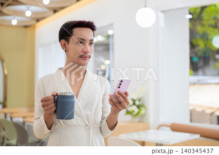 Non-binary gay Thai man looking away while holding smartphone and coffee mug in modern cafe Non-binary gay Thai man looking away while holding smartphone and coffee mug in modern cafe 140345437
