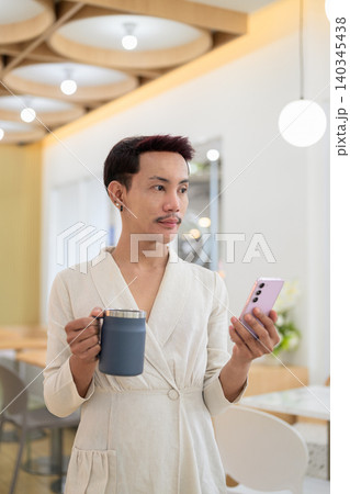 Vertical portrait of non-binary gay Thai man looking away while holding smartphone and coffee mug Vertical portrait of non-binary gay Thai man looking away while holding smartphone and coffee mug 140345438