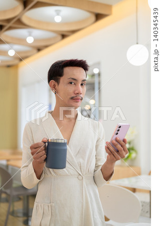 Vertical portrait of non-binary gay Thai man looking at camera while holding smartphone and coffee mug Vertical portrait of non-binary gay Thai man looking at camera while holding smartphone and coffee mug 140345439