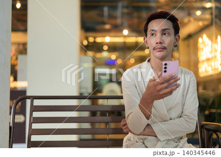 Non-binary gay Thai man looking away while holding smartphone on outdoor bench at night 140345446