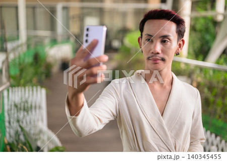 Non-binary gay Thai man looking at smartphone screen taking selfie on outdoor patio walkway Non-binary gay Thai man looking at smartphone screen taking selfie on outdoor patio walkway 140345455