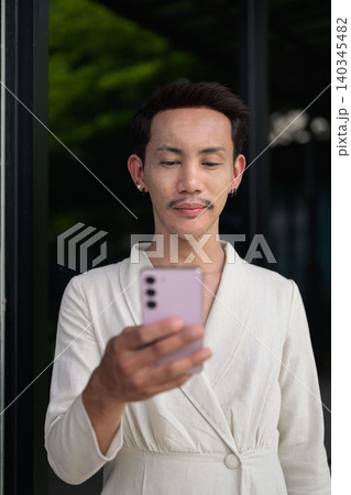 Vertical portrait of non-binary gay Thai man using smartphone outside modern storefront Vertical portrait of non-binary gay Thai man using smartphone outside modern storefront 140345482