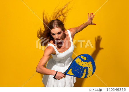 Dynamic female padel player reaching forward with racket in yellow studio. 140346256