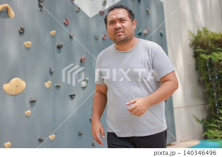 Portrait of a determined overweight Asian man standing by an outdoor rock climbing wall. Serious chubby male resting after bouldering exercise. Active fitness and weight loss journey concept. 140346496