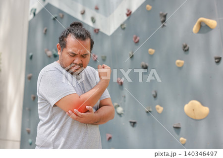 Overweight Asian man suffering from elbow joint pain with glowing red graphic overlay after rock climbing exercise. Sports injury, muscle inflammation and healthcare concept near bouldering wall. 140346497