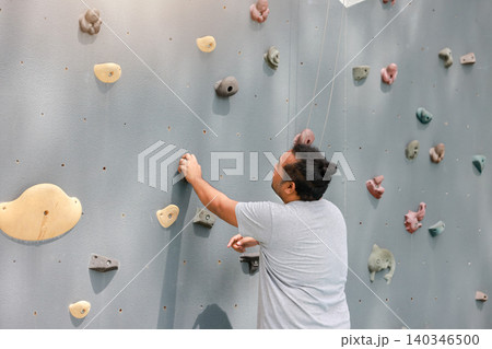 Back view of a determined Asian man reaching for a hold on an artificial rock climbing wall. Outdoor bouldering exercise, active fitness training and reaching goal concept. Back view of a determined Asian man reaching for a hold on an artificial rock climbing wall. Outdoor bouldering exercise, active fitness training and reaching goal concept. 140346500