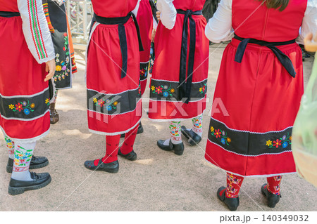 Close view of Bulgarian women wearing red traditional folk costumes with floral embroidery, outdoor cultural event. Heritage clothing, patterned socks, festive folk tradition, ethnic cultural heritage 140349032