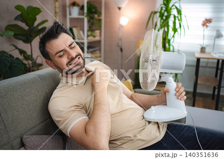 Man suffering from summer heat holding electric fan at home Man suffering from summer heat holding electric fan at home 140351636
