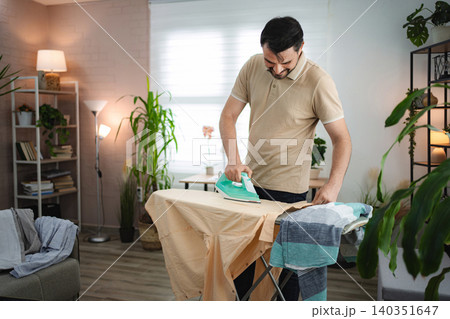 Man diligently ironing clothes on ironing board in living room 140351647