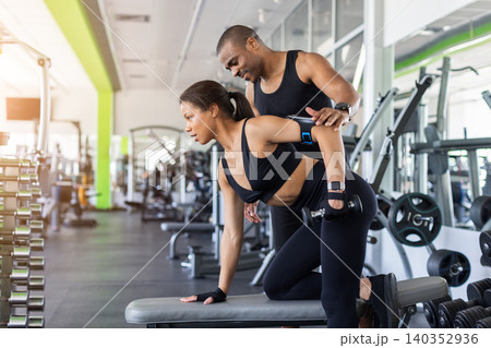 Personal trainer coaching woman during one-arm dumbbell row exercise at modern gym Personal trainer coaching woman during one-arm dumbbell row exercise at modern gym 140352936