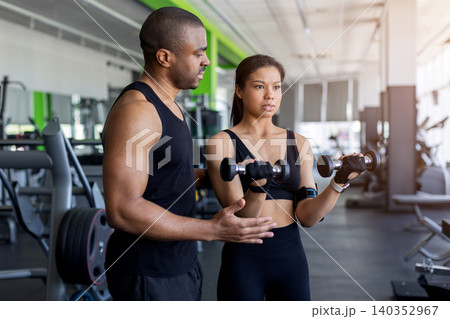Personal trainer coaching young woman performing dumbbell curls in modern fitness club Personal trainer coaching young woman performing dumbbell curls in modern fitness club 140352967