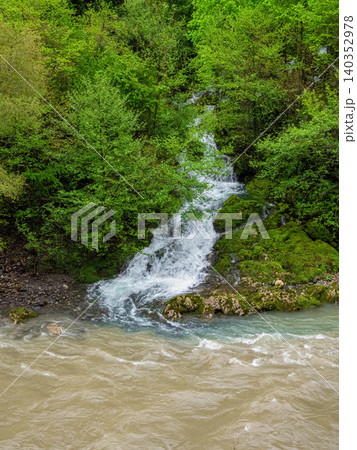 A small waterfall plunges over mossy rocks into a muddy, stormy river after rains. 140352978