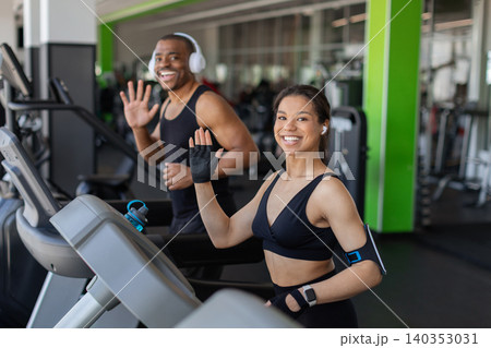 Smiling young black waving while exercising on treadmills in modern gym Smiling young black waving while exercising on treadmills in modern gym 140353031