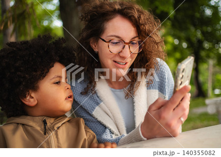 Mother and child bonding over smartphone outdoors in park Mother and child bonding over smartphone outdoors in park 140355082