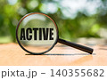 Magnifying glass with text ACTIVE on wooden table and green background. 140355682