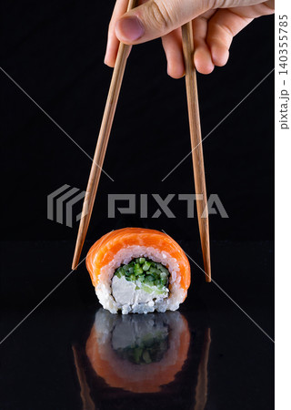 Chopsticks holding a single california uramaki roll with orange tobiko caviar over black reflection Chopsticks holding a single california uramaki roll with orange tobiko caviar over black reflection 140355785