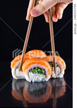Chopsticks lifting a philadelphia sushi roll from a line on dark glossy background Chopsticks lifting a philadelphia sushi roll from a line on dark glossy background 140355795