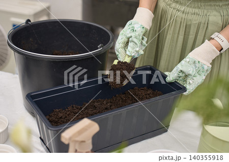 Woman fills black planter box with potting soil using a hand trowel at outdoor garden table Woman fills black planter box with potting soil using a hand trowel at outdoor garden table 140355918