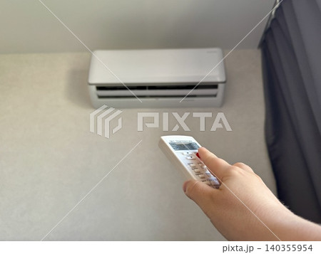 Close-up female hand holding white remote control pointing at air conditioner, bright modern apartment interior, climate control and home comfort concept 140355954