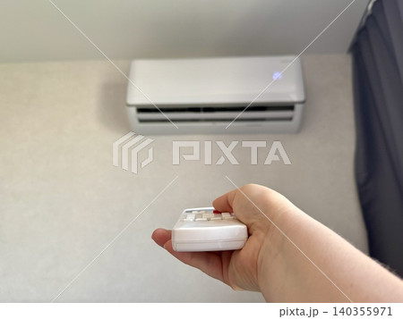 Close-up female hand holding white remote control pointing at air conditioner, bright modern apartment interior, climate control and home comfort concept 140355971