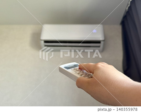 Close-up female hand holding white remote control pointing at air conditioner, bright modern apartment interior, climate control and home comfort concept Close-up female hand holding white remote control pointing at air conditioner, bright modern apartment interior, climate control and home comfort concept 140355978