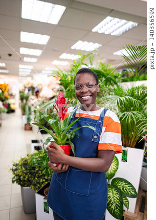 Young florist holding a red potted plant smiling at the camera in a flower shop 140356599