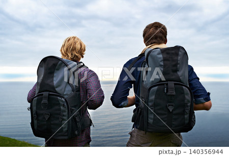 Couple, backpack and hiking by ocean for travel, sightseeing or outdoor journey in nature. Rear view of man, woman or hikers carrying bag on back for trekking, fitness or adventure with cloudy sky 140356944