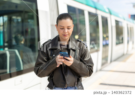Young woman waiting for tram at stop with mobile phone Young woman waiting for tram at stop with mobile phone 140362372