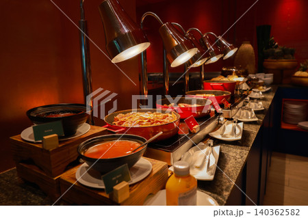 Luxury hotel breakfast buffet hot food catering station featuring crispy bacon, hot sausages, and tasty sauces in red pans under warm heating lamps. Morning meal service setup for guest dining. Luxury hotel breakfast buffet hot food catering station featuring crispy bacon, hot sausages, and tasty sauces in red pans under warm heating lamps. Morning meal service setup for guest dining. 140362582