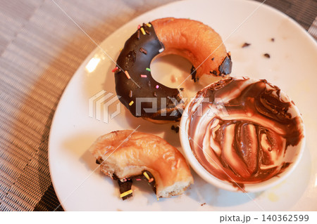Partially eaten chocolate frosted donut with colorful sprinkles and a messy empty small bowl of chocolate cream spread on a white plate. Sweet leftover dessert food after breakfast meal at a cafe. Partially eaten chocolate frosted donut with colorful sprinkles and a messy empty small bowl of chocolate cream spread on a white plate. Sweet leftover dessert food after breakfast meal at a cafe. 140362599