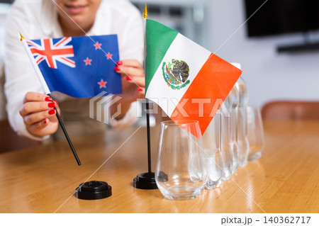 A woman in a white blouse with friendly smile arrangs flags of Mexico and New Zealand for meeting. A woman in a white blouse with friendly smile arrangs flags of Mexico and New Zealand for meeting. 140362717