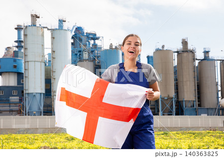 near suburban factory on sunny day, stands gleeful female worker in overalls and holds English flag 140363825