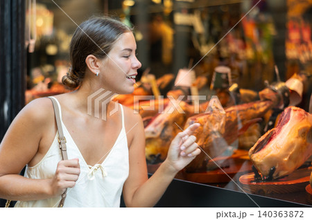 Passerby young woman examines smoked whole meat pork leg in shop window. Passerby young woman examines smoked whole meat pork leg in shop window. 140363872